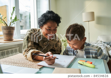 Woman wearing military outfit sitting at the table with her little son and drawing 84534616