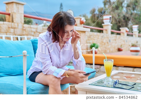 Sad upset mature woman sitting outdoors at table in cafe with smartphone Sad upset mature woman sitting outdoors at table in cafe with smartphone 84534862
