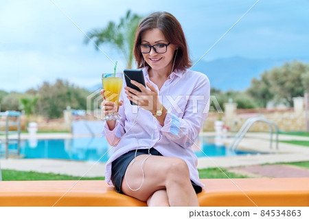Middle-aged woman relaxing near pool using headphones and smartphone Middle-aged woman relaxing near pool using headphones and smartphone 84534863
