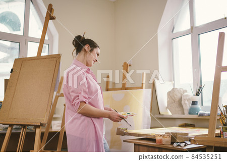 Cute woman paints on canvas in an art workshop. Artist creating picture. Art school or studio. Work with paints, brushes and easel. Hobby and leisure concept. Woman painter at workspace 84535251