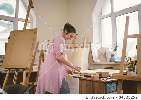 Caucasian woman artist working on a painting in bright daylight studio. Happy artist draws an art project with paints and a brush in the workshop. Hobby. Artist at work. Creative profession Caucasian woman artist working on a painting in bright daylight studio. Happy artist draws an art project with paints and a brush in the workshop. Hobby. Artist at work. Creative profession 84535252