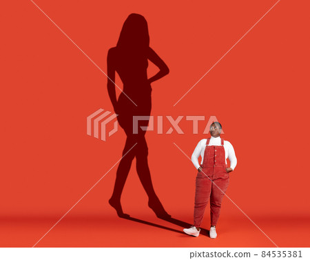 Conceptual image with fat woman standing near wall with shadow of slim fashion model isolated on red background Conceptual image with fat woman standing near wall with shadow of slim fashion model isolated on red background 84535381