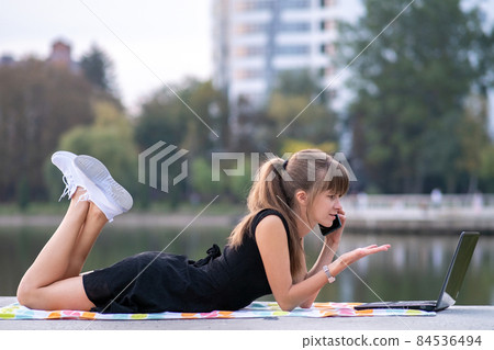 Young woman working behind laptop while talking on mobile phone lying down in summer park outdoors. 84536494