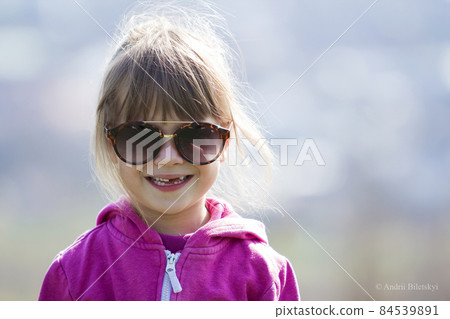 Portrait of cute pretty little blond preschool girl in pink sweater and dark sunglasses smiling happily in camera with funny child toothless smile against blurred light blue background outdoors. 84539891