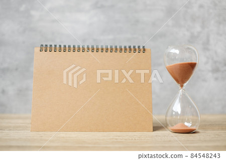 Hourglass with Blank notebook on table, Sand flowing through the bulb of Sandglass measuring the passing. countdown, deadline, Life time and Retirement concept 84548243