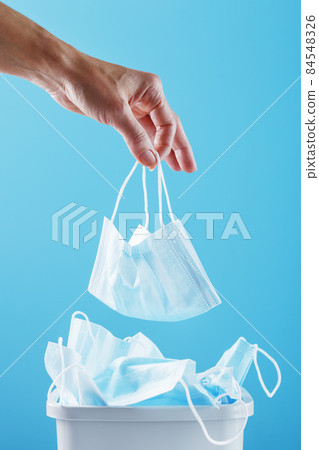 Hand throws a used face shield into the trash can on a light blue background. Hand throws a used face shield into the trash can on a light blue background. 84548326