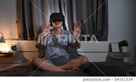 Exited asian man wearing virtual reality headset playing video game or watching movie in bedroom. Exited asian man wearing virtual reality headset playing video game or watching movie in bedroom. 84551334