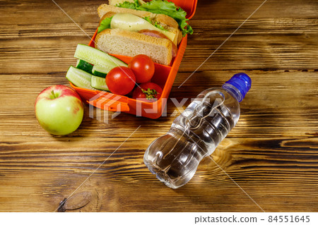 Bottle of water, apple and lunch box with sandwiches and fresh vegetables on a wooden table Bottle of water, apple and lunch box with sandwiches and fresh vegetables on a wooden table 84551645