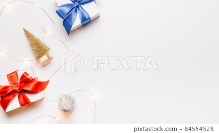 Christmas gift. White gift with red bow, gold balls and sparkling lights garland in xmas decoration on white background for greeting card. Xmas backdrop with space for text. Christmas gift. White gift with red bow, gold balls and sparkling lights garland in xmas decoration on white background for greeting card. Xmas backdrop with space for text. 84554528