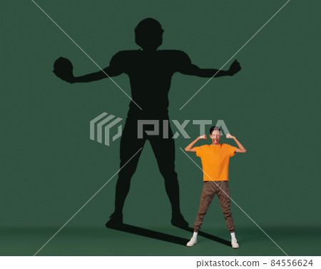 Conceptual image with young boy dreaming about future sport career and shadow of fit male football player on dark green background 84556624