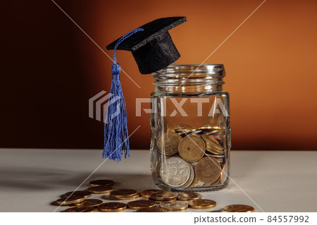 Jar with coins and graduation hat on a table, scholarship and savings concept Jar with coins and graduation hat on a table, scholarship and savings concept 84557992