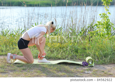 Young beautiful girl sat down on knee and puts weighting on foot for exercise outdoors in woods on background of river. 84562922
