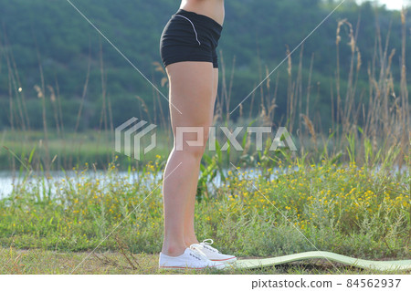 Photo slender legs of girl in shorts and sneakers in woods near mat against background of river. 84562937