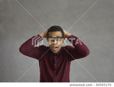 Portrait of a young dark-skinned man who grabs his head with a surprised expression on his face. Shocked emotional man simply cannot believe what he has heard or seen. Gray background. 84565270