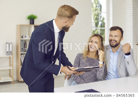 Young family is sincerely happy and celebrates signing of contract of sale or approval of loan. Caucasian couple happily clenching their fists and shouting cheers looking at realtor or bank manager. Young family is sincerely happy and celebrates signing of contract of sale or approval of loan. Caucasian couple happily clenching their fists and shouting cheers looking at realtor or bank manager. 84565365