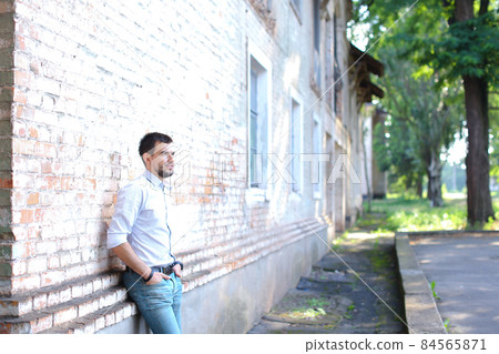 Young handsome guy standing near wall, looking ahead of itself and holds hands in pockets of pants. 84565871