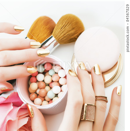 woman hands with golden manicure and many rings holding brushes, cosmetic and rose flower on white background, spa treatment concept 84567829