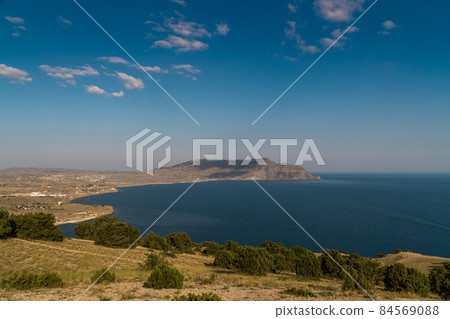 The Republic of Crimea. July 18, 2021. Picturesque view in clear sunny weather of the cape and Mount Meganom from the height of Mount Alchak in Sudak. The Republic of Crimea. July 18, 2021. Picturesque view in clear sunny weather of the cape and Mount Meganom from the height of Mount Alchak in Sudak. 84569088