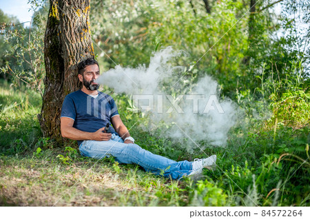 Bearded vaper smoking an electronic smoke device in nature. Electronic cigarette. 84572264