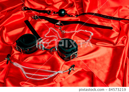 Leather collar with metal chain leash, gag ball, handcuffs, nipple clamps on a red satin sheet. Leather collar with metal chain leash, gag ball, handcuffs, nipple clamps on a red satin sheet. 84573219
