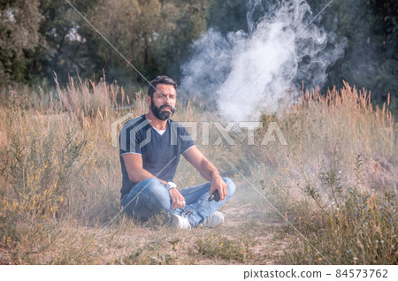 Brutal vaper enjoying an electronic cigarette on the forest ground. Hybrid cigarette. 84573762