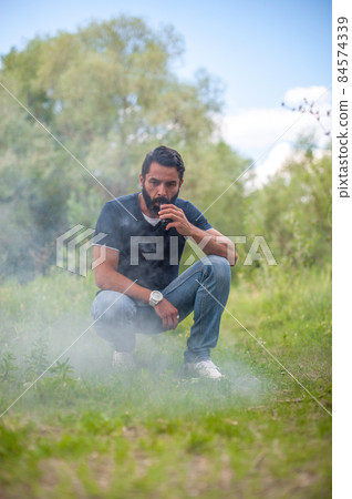 Handsome vaper in the fresh air smoking an electronic cigarette. Non-tobacco smoking. Handsome vaper in the fresh air smoking an electronic cigarette. Non-tobacco smoking. 84574339