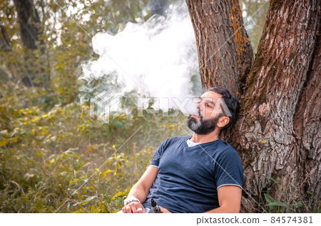Bearded vaper smoking an electronic cigarette outdoorr. Smoking cessation tobacco. Bearded vaper smoking an electronic cigarette outdoorr. Smoking cessation tobacco. 84574381
