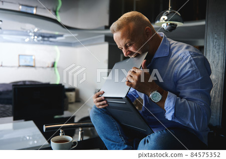 Middle aged angry Caucasian man, entrepreneur in casual clothes sitting on a table with a cup of coffee and holding a laptop in his hands, biting it with rage Middle aged angry Caucasian man, entrepreneur in casual clothes sitting on a table with a cup of coffee and holding a laptop in his hands, biting it with rage 84575352