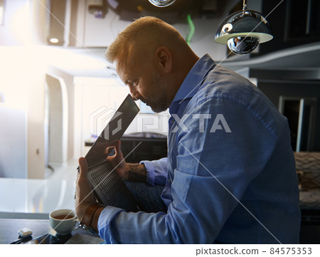 Side portrait to a happy successful middle aged businessman holding laptop near his face, sitting on a table with cup of coffee against the background of sunbeams falling through the window at home Side portrait to a happy successful middle aged businessman holding laptop near his face, sitting on a table with cup of coffee against the background of sunbeams falling through the window at home 84575353