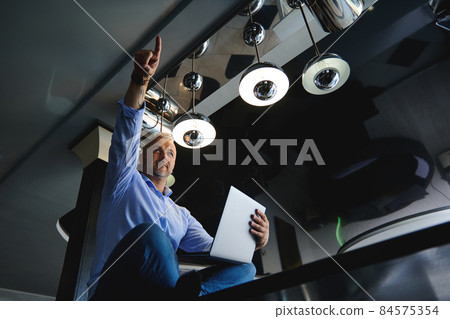 View from the bottom to a successful middle aged European man in casual denim jeans and blue shirt with laptop, pointing finger up, sitting on a table at home kitchen, working remotely from home View from the bottom to a successful middle aged European man in casual denim jeans and blue shirt with laptop, pointing finger up, sitting on a table at home kitchen, working remotely from home 84575354