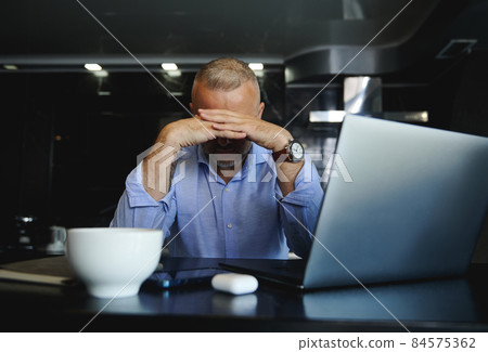 Disappointed middle-aged businessman sitting at a desk with a laptop, looking down thoughtfully hiding his face with his hands 84575362