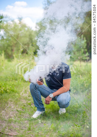Modern man enjoying an e-cigarette on the forest floor. The man smokes. Modern man enjoying an e-cigarette on the forest floor. The man smokes. 84575884