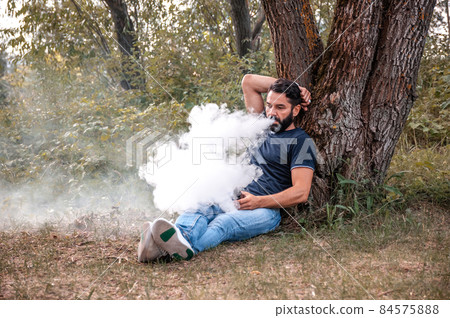 Stylish bearded smoker blows up a couple an electronic cigarette outside. Modern ways of quitting tobacco. Stylish bearded smoker blows up a couple an electronic cigarette outside. Modern ways of quitting tobacco. 84575888