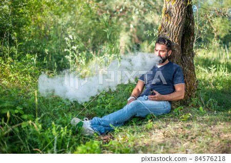 Stylish brutal vaper smoking an electronic smoke device on the forest ground. Alternative to smoking. Stylish brutal vaper smoking an electronic smoke device on the forest ground. Alternative to smoking. 84576218