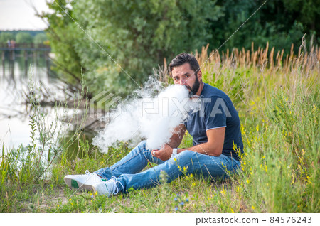 Stylish brutal smoker smoking an electronic cigarette in the daylight. Non-tobacco smoking. Stylish brutal smoker smoking an electronic cigarette in the daylight. Non-tobacco smoking. 84576243