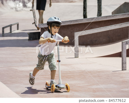 Little boy rides kick scooter in skate park. Special concrete bowl structures in urban park. Training to skate at summer. Little boy rides kick scooter in skate park. Special concrete bowl structures in urban park. Training to skate at summer. 84577612