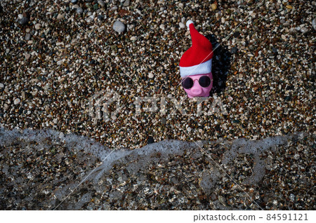 Piggy bank in a hat of santa claus and sunglasses on a pebble beach by the sea. Piggy bank in a hat of santa claus and sunglasses on a pebble beach by the sea. 84591121