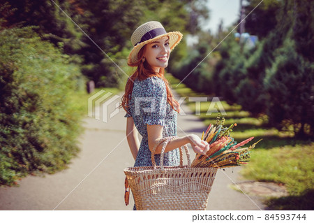 back view of young smiling adult girl with stylish straw bag and straw hat who walks in park outdoor 84593744