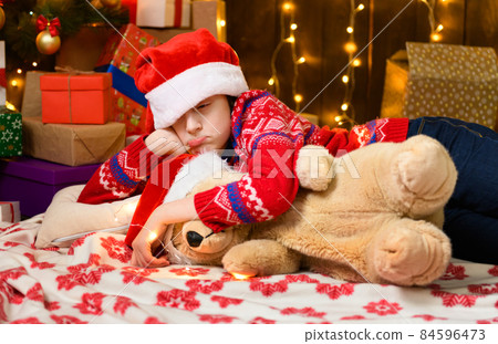 Child girl posing and dreaming in new year or christmas decoration. Holiday lights and gifts, Christmas tree decorated with toys. She's wearing a red sweater and a Santa helper hat Child girl posing and dreaming in new year or christmas decoration. Holiday lights and gifts, Christmas tree decorated with toys. She's wearing a red sweater and a Santa helper hat 84596473