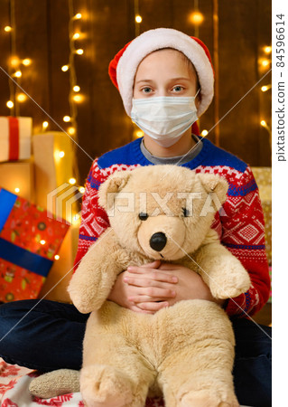Child girl as Santa helper posing in new year decoration. Wearing a protective face mask against viruses, coronaviruses. She's playing with a toy bear. Holiday lights and lots of gifts, an elegant 84596614