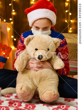 Child girl as Santa helper posing in new year decoration. Wearing a protective face mask against viruses, coronaviruses. She's playing with a toy bear. Holiday lights and lots of gifts, an elegant 84596618