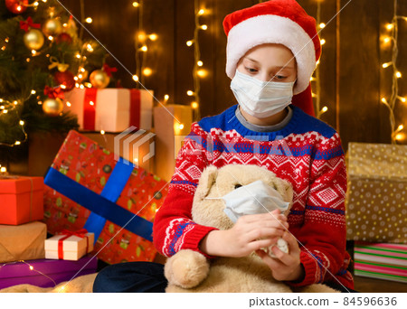 Child girl as Santa helper posing in new year decoration. Wearing a protective face mask against viruses, coronaviruses. She plays with a toy bear, cures him and gives him pills. 84596636