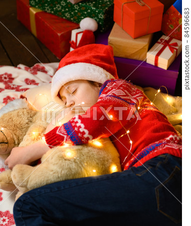 Child girl sleeping in new year or christmas decoration. Holiday lights and gifts, Christmas tree decorated with toys. She's wearing a red sweater and a Santa helper hat Child girl sleeping in new year or christmas decoration. Holiday lights and gifts, Christmas tree decorated with toys. She's wearing a red sweater and a Santa helper hat 84596683