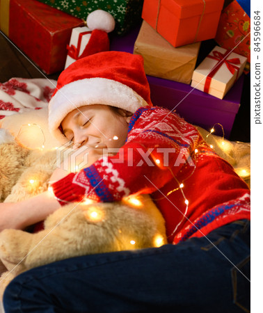 Child girl sleeping in new year or christmas decoration. Holiday lights and gifts, Christmas tree decorated with toys. She's wearing a red sweater and a Santa helper hat Child girl sleeping in new year or christmas decoration. Holiday lights and gifts, Christmas tree decorated with toys. She's wearing a red sweater and a Santa helper hat 84596684