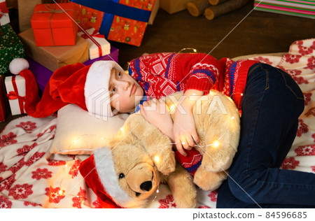 Child girl sleeping in new year or christmas decoration. Holiday lights and gifts, Christmas tree decorated with toys. She's wearing a red sweater and a Santa helper hat Child girl sleeping in new year or christmas decoration. Holiday lights and gifts, Christmas tree decorated with toys. She's wearing a red sweater and a Santa helper hat 84596685