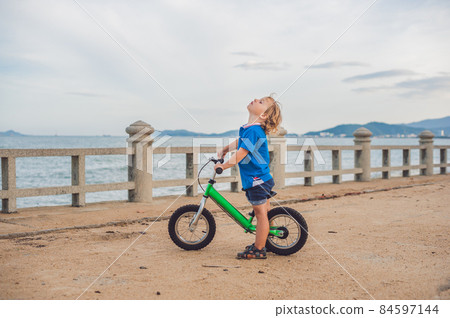 Active blond kid boy driving bicycle in the park near the sea. Toddler child dreaming and having fun on warm summer day. outdoors games for children 84597144