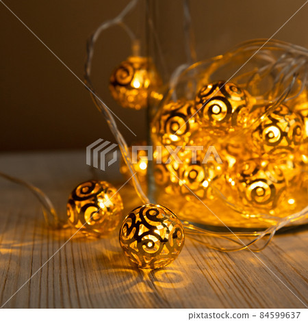New Year's garland. Golden metal balls in glass jar on light wooden table background. Christmas yellow gold garland lights from LED bulbs. Front view. Square photo. Selective focus. 84599637
