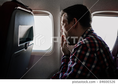 a man in a plaid shirt sits by the window of the plane and yawns. long flight.  84600326