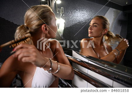 Charming young Caucasian blonde woman in swimsuit combing her blond hair and looking at her reflection in bathroom mirror 84601858