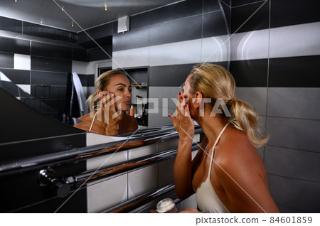 Charming young Caucasian blonde woman in swimsuit applying moisturizer, performing smoothing rejuvenating facial massage looking at her reflection in bathroom mirror 84601859